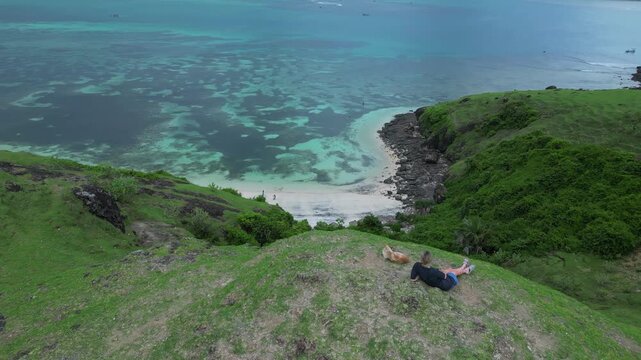 Woman Relaxing With Her Pet. Woman And Her Dog Observe Peaceful Bay From Hillside. Caucasian Woman And Her Canine Friend Gaze Over Tranquil Shoreline From Grassy Hilltop,Lombok