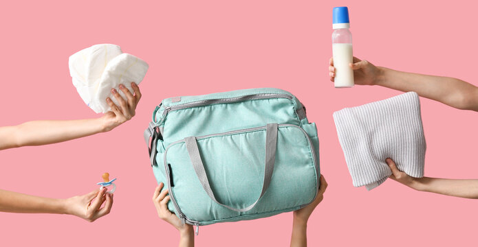 Hands holding baby essentials and diaper bag on pink background