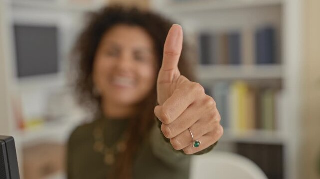 Woman smiling and giving thumbs up with hand and green ring at a studio desk with computer monitor and bookshelf; confidence success.