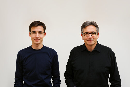close up portrait of a young man and a mature man with grey hair standing together side by side both wearing glasses 