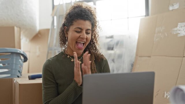 Woman clapping hands and smiling at laptop amid moving boxes in building; joy relief accomplishment.