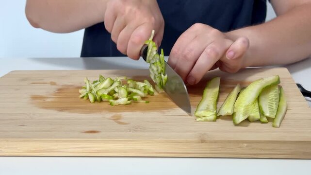 Woman using knife to thinly slice cucumber on cutting board in kitchen, preparing meal