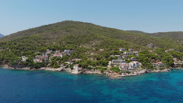 Slow panoramic drone shot moving backward from coastal hotels on a pine-covered hill, revealing Skala village on a sunny day. Agistri island, Greece