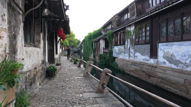 A narrow cobblestone path lined with traditional buildings and red lanterns runs alongside Zhouzhuang's historic canal. Ideal for travel documentaries and cultural features.