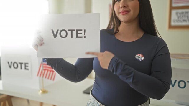 Woman holding paper sign reading vote, pointing finger to sign, wearing voted sticker on chest inside a public building; civic duty pride.