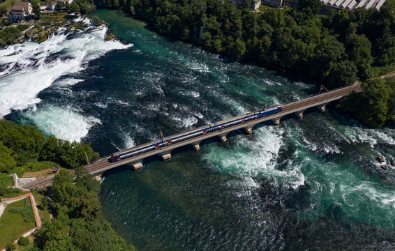 Aerial view of a train crossing the stone bridge over the rushing waters of the Rhine Falls surrounded by lush green trees in Schaffhausen, Schaffhausen, Switzerland.
