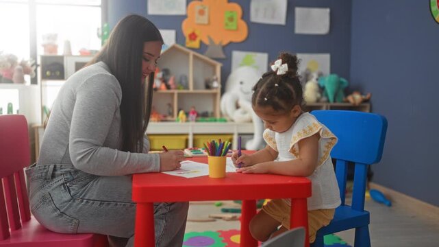 Teacher guides little girl drawing with crayons at a red table in a preschool building; nurturing learning moment.