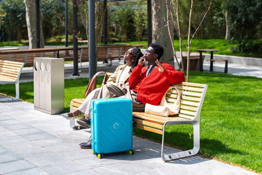 Carefree african american tourists couple man and woman enjoying rest while sitting in green square, black male and female partners travelers relax on bench in park sharing impressions about trip.