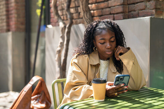 Thoughtful african american female on coffee break in cafe, sitting at table scrolling social media on smartphone, relaxed black female reading news on phone, enjoys sunny weather and peaceful time.