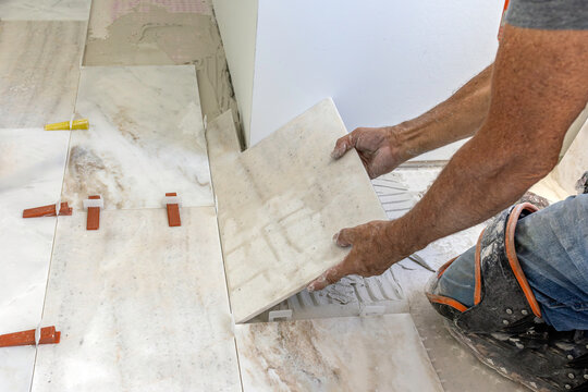 Side view of installer hands carefully laying a large marble tile on a notched mortar bed with a leveling system during floor installation