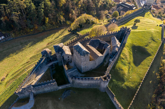Aerial view of Castello di Montebello, a historic medieval fortress with stone walls and towers, surrounded by green grassy slopes and autumn trees in Bellinzona, Ticino, Switzerland.