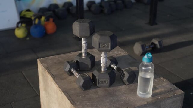 Hex dumbbells and a water bottle arranged on a wooden plyometric box in a modern indoor gym with smooth lateral camera movement.