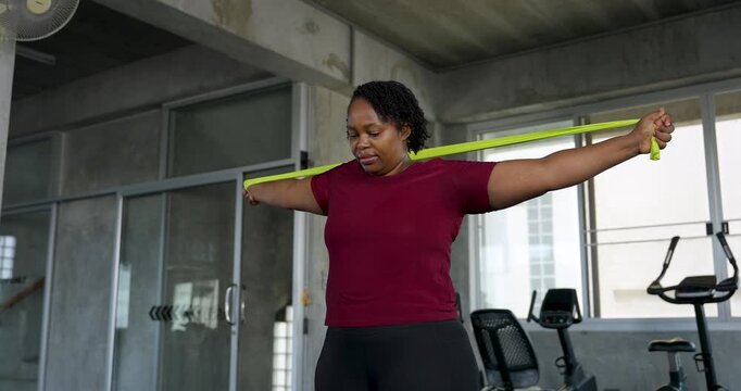 African American plus size woman moves resistance band behind shoulder and overhead during mobility exercise in modern gym. Curvy female athlete builds strength, wellness, body confidence routine.