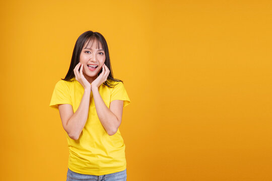 Surprised young Asian woman in a bright yellow t-shirt, looking at camera with open mouth, smiling widely, hands on cheeks disbelief. Cheerful studio portrait with empty yellow background copy space.