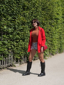 Young Asian woman wearing a red jacket and black leather boots stands on a gravel pathway next to a green hedge while smiling