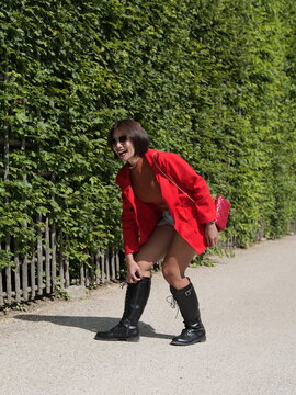 Young Asian woman wearing a red jacket and black leather boots bending down to adjust her boot on a gravel pathway next to a green hedge
