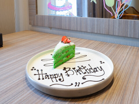 ​Pandan klepon cake slice with happy birthday chocolate writing on a white plate, celebration dessert serving close up