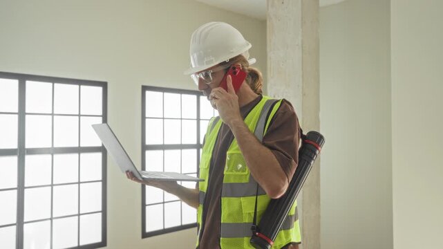 Man architect in hard hat and reflective vest holds laptop in left hand and phone to ear while checking plans inside a building; communication coordination focus.