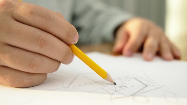 A close-up of a man's hand holding a yellow pencil and drawing or sketching on white paper on a desk.