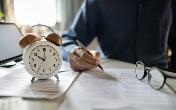 close up alarm clock 10 AM with asian businessman working on desk with laptop reviewing document, professional lawyer checking financial report, business contract