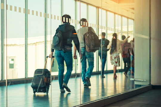 Photograph through large clear glass wall with blurred image of departing passengers and luggage walking airplane boarding corridor from terminal to plane in background. Selective focus on glass.