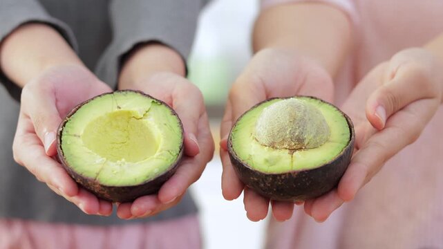 Two Asian friends showing fresh cut avocado halves in the kitchen during healthy green juice preparation, perfect for clean eating, weight loss, and nutritious diet concepts.