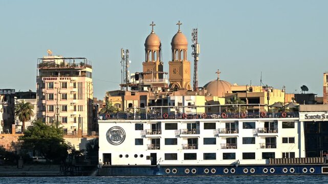 A view from the water showing a multi-deck Nile cruise liner docked at the Luxor embankment, with the prominent twin towers and dome of a Christian church rising immediately behind the urban skyline.