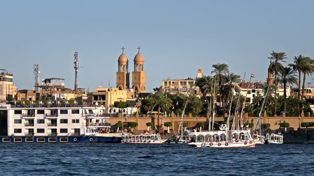 A golden hour view from the water focusing on a large multi-deck Nile cruise boat docked along the Luxor waterfront, with the prominent twin towers and dome of a church rising directly behind it again