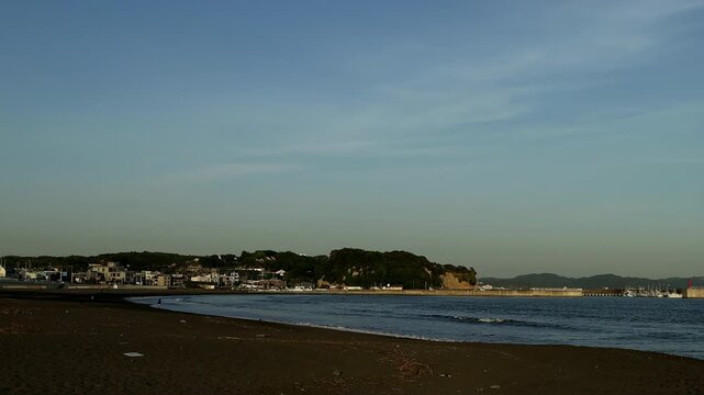 Calm evening coastline in Kamakura, Japan, featuring soft waves rolling onto the sandy beach and a distant island on the horizon. Japanese seaside travel and nature background.