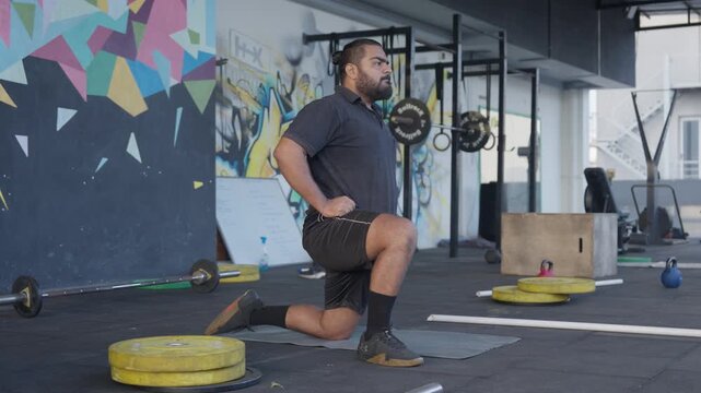 Bearded male athlete performing a half kneeling hip flexor stretch during a warm up session in a modern indoor fitness studio with a city view