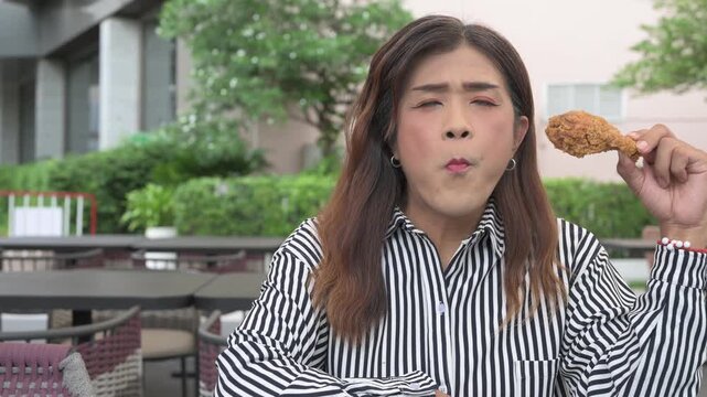 Happy Thai Isan transgender woman in 40s eating crispy fried chicken outdoors. Horizontal medium shot portrait of a mature non-binary Southeast Asian trans woman eating.