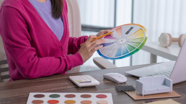 A close up of a designer's hand holding a pencil and rotating a colorful color wheel chart at her desk with a laptop nearby.