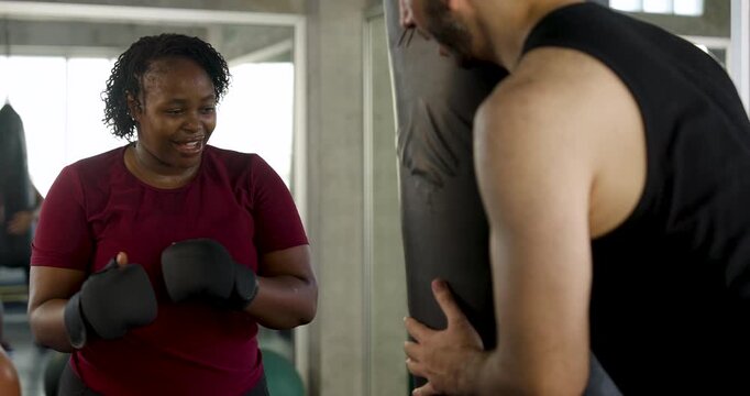 Plus size African American woman smiles, resets stance, then throws punches at heavy bag while male trainer supports session in modern gym, reflecting boxing progress, cardio effort, strength.