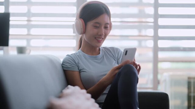 Asian woman in sports clothing using smartphone with headphones at home