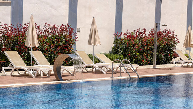 Luxury outdoor swimming pool with empty sun loungers, umbrellas and cascading water feature at a modern hotel resort. Summer vacation, relaxation, wellness and premium travel destination.
