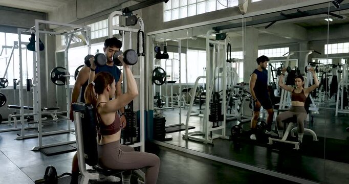 Young Asian couple practicing teamwork and wellness routine in modern gym, seated woman lifting dumbbell while man coaches form, partner workout support and healthy lifestyle motivation goals.