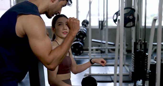 Young Asian couple resting between sets in modern gym, man explaining arm exercise form to woman holding dumbbell, partner coaching with care, support, recovery, fitness motivation, wellness.