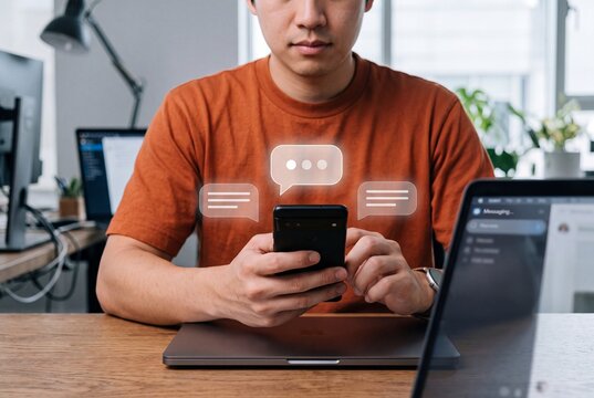 Young man using a mobile phone to send instant messages in a bright office environment with digital speech bubbles floating above the device display