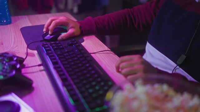 Man clicking mouse with hand at keyboard, wearing hoodie with headset cable, popcorn bowl on desk in studio; concentration leisure.