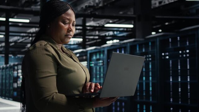 Software developer uses a laptop computer in a server hub for technology workflow management, supporting equipment monitoring, system reliability and server infrastructure management.