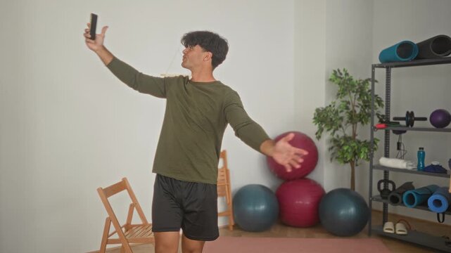 Man holds smartphone aloft with outstretched hand for a selfie during yoga training in a fitness studio, wearing olive long sleeve shirt and black shorts; mindful focus.