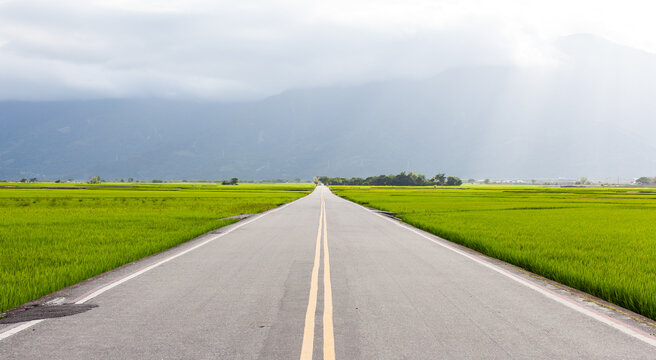 A straight road runs through vibrant green rice fields. Distant mountains frame the horizon under a cloudy sky. Chishang's rural landscape showcases lush farmland and serene countryside.