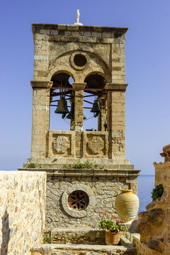 A stone bell tower with bells and a cross on top stands against a clear blue sky, with the sea visible in the background.