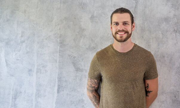 Portrait of bearded caucasian man with tattoos standing against a textured gray background, expressing confidence, friendliness, and a casual modern lifestyle.