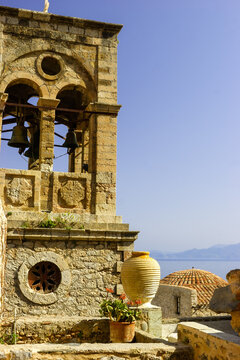 A bell tower with bells and a large amphora stand in front of a tiled dome and the sea in Monemvasia, Greece.