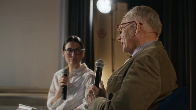 Medium shot of senior Caucasian man speaking into microphone during book presentation with adult woman listening. Perfect for educational seminars or literary event promotions
