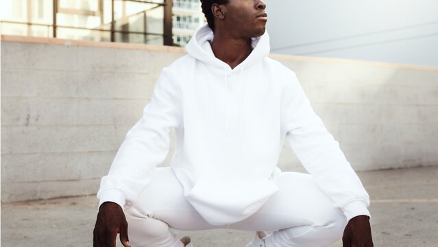 Person in a white hoodie and pants squatting outdoors, urban setting.