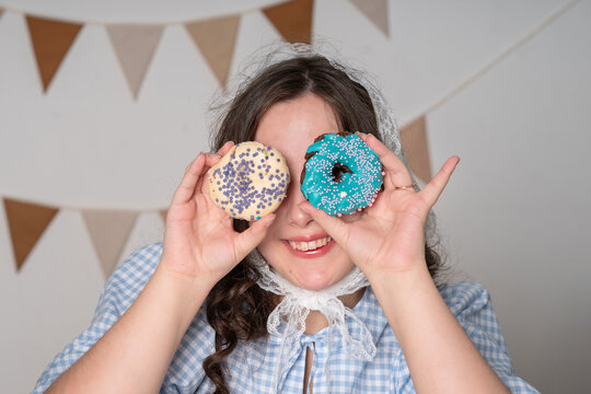A beautiful girl in a lace headscarf and a blue checkered blouse holds a donut in her hands. The girl holds donuts near her eyes like glasses