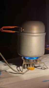 A portable lightweight gas camping burner stove with a bright blue flame cooking food in a grey anodized aluminum pot with an orange handle. Outdoor survival tourist equipment setup