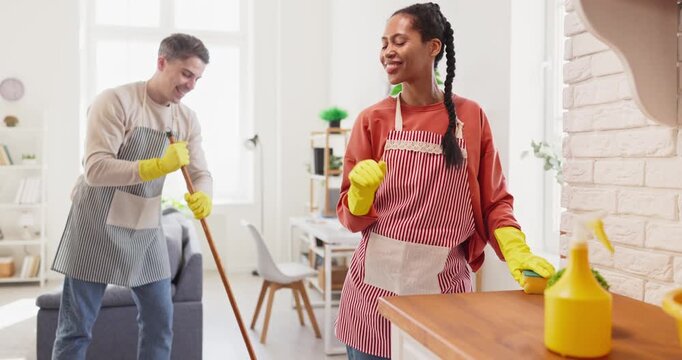 Couple cleaning home teamwork highfive man woman. They celebrate finished chores in a bright apartment as the man holds a mop and both wear aprons and gloves. Clear team success unity concept.
